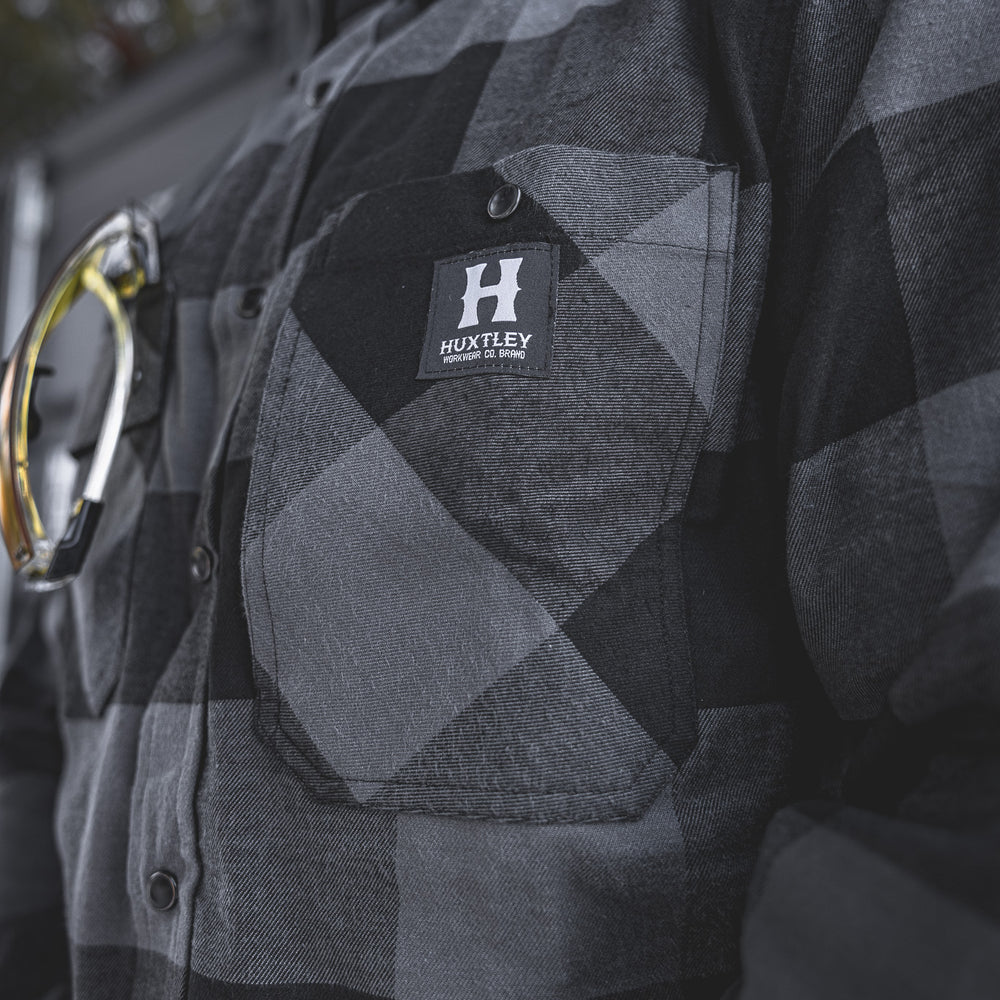 
                  
                    Up-close view of the Huxtley HD-57 Flex Flannel in Charcoal/Black, highlighting the chest pocket stitching and iconic Huxtley Workwear logo. Built for tradesmen who take pride in their gear.
                  
                