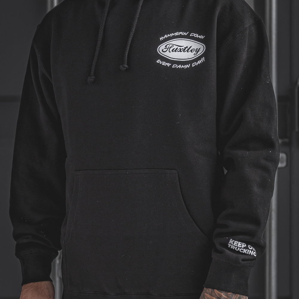 Front view of the black Huxtley Keep On Trucking hoodie. Features chest print with “Hammerin’ Down Every Damn Day!!” and logo hit on the sleeve—built for the cold grind.