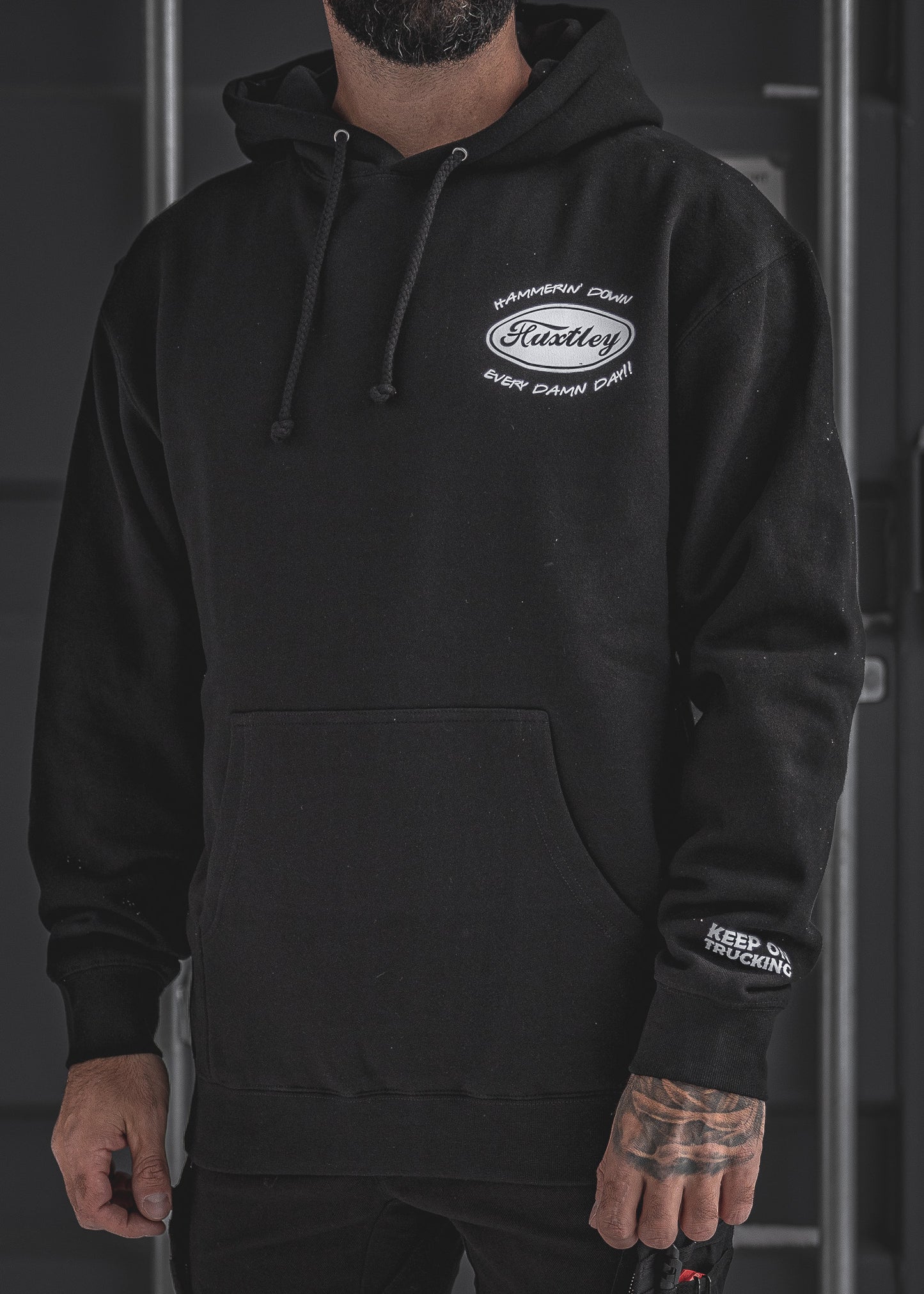 Front view of the black Huxtley Keep On Trucking hoodie. Features chest print with “Hammerin’ Down Every Damn Day!!” and logo hit on the sleeve—built for the cold grind.
