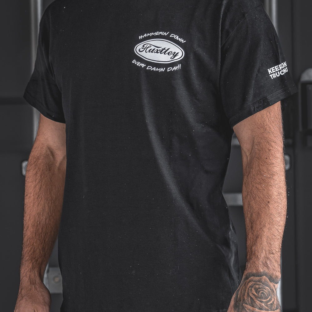 Front view of the Huxtley Keep On Trucking Tee in black, featuring the “Hammerin’ Down Every Damn Day!” chest logo and sleeve print. A bold statement for those who don’t take their foot off the gas.