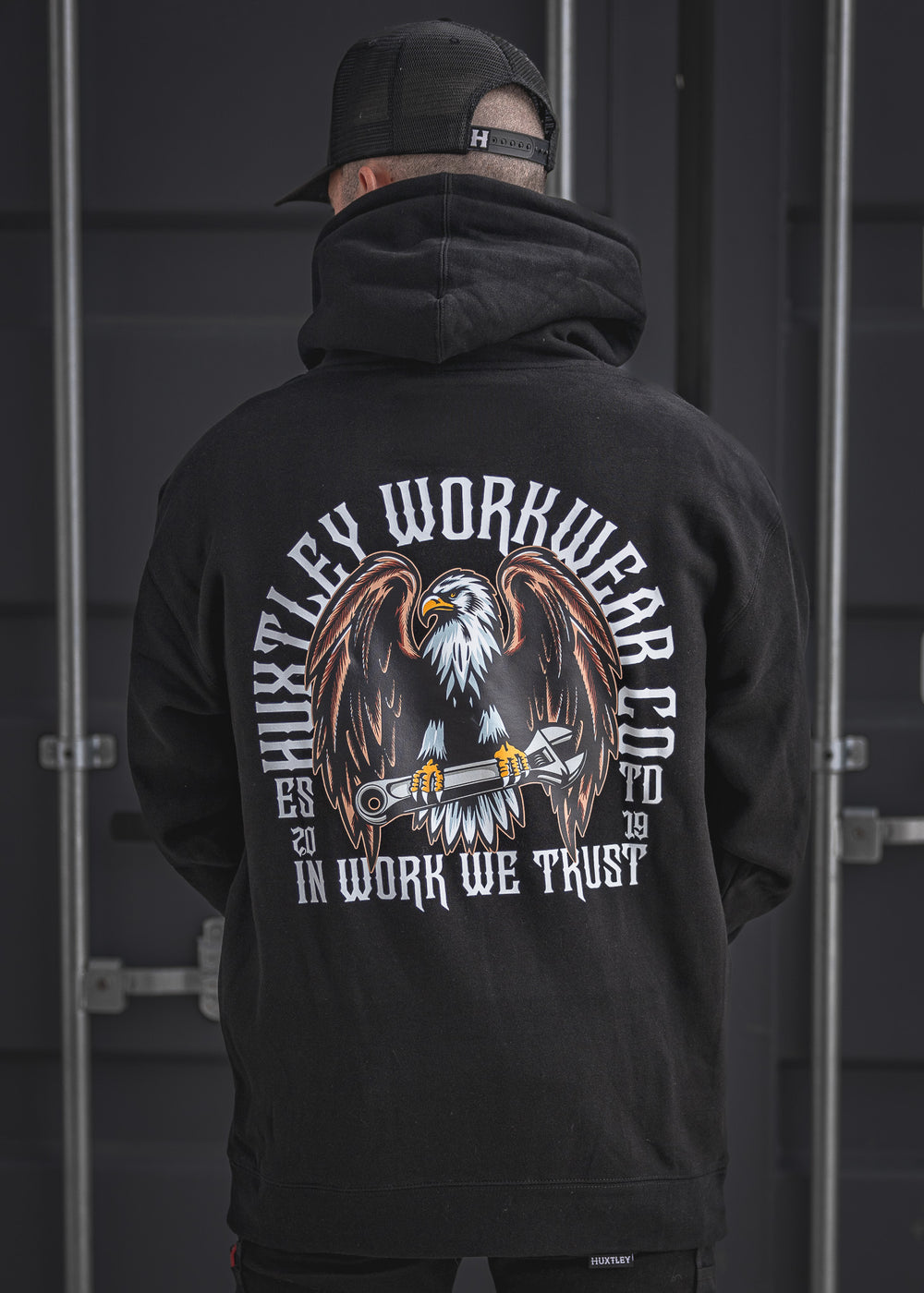 Back view of the In Work We Trust Hoodie in black, featuring a bold eagle clutching a wrench and vintage-styled Huxtley Workwear Co. branding. Built for the site and proud of it.