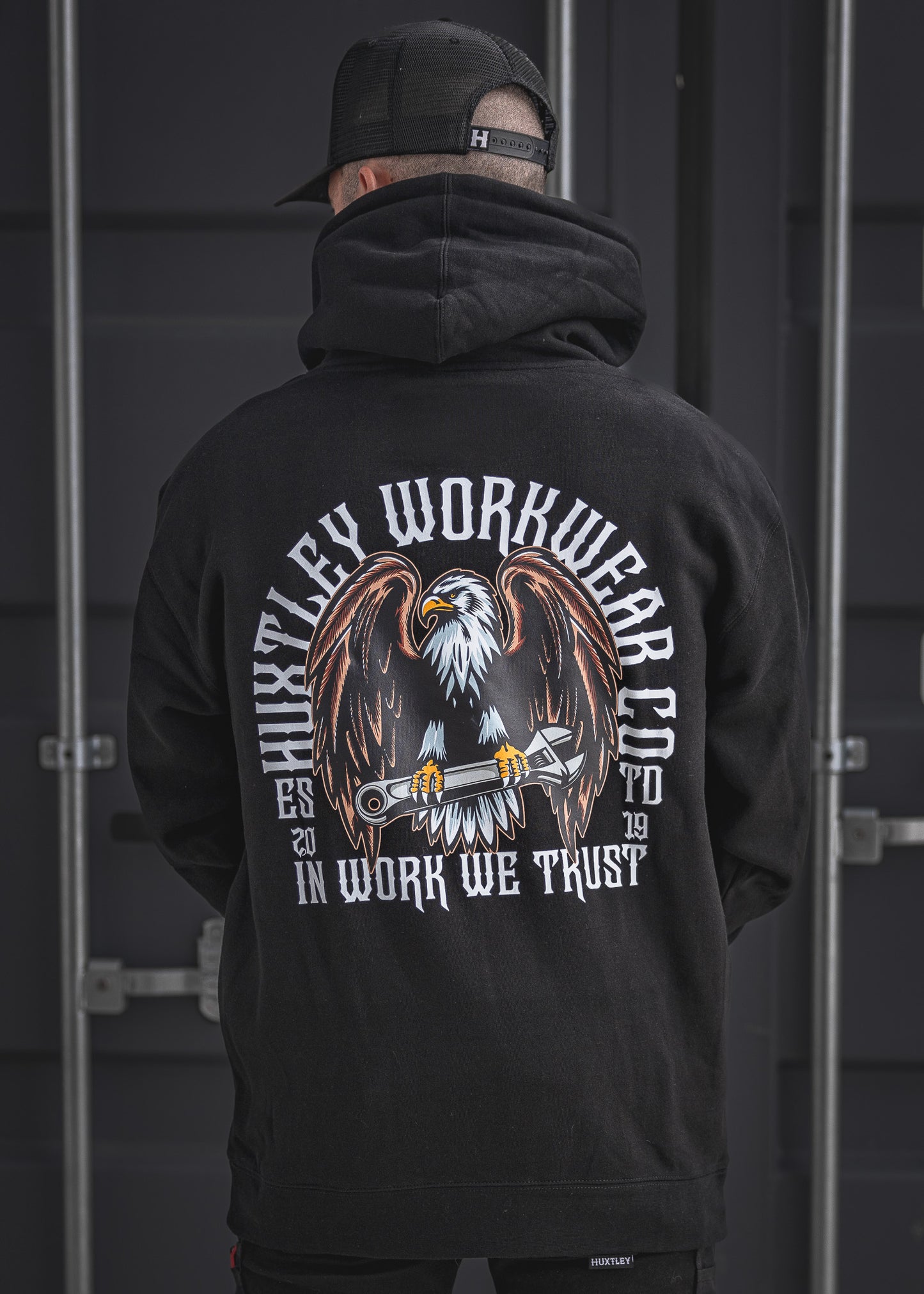 Back view of the In Work We Trust Hoodie in black, featuring a bold eagle clutching a wrench and vintage-styled Huxtley Workwear Co. branding. Built for the site and proud of it.
