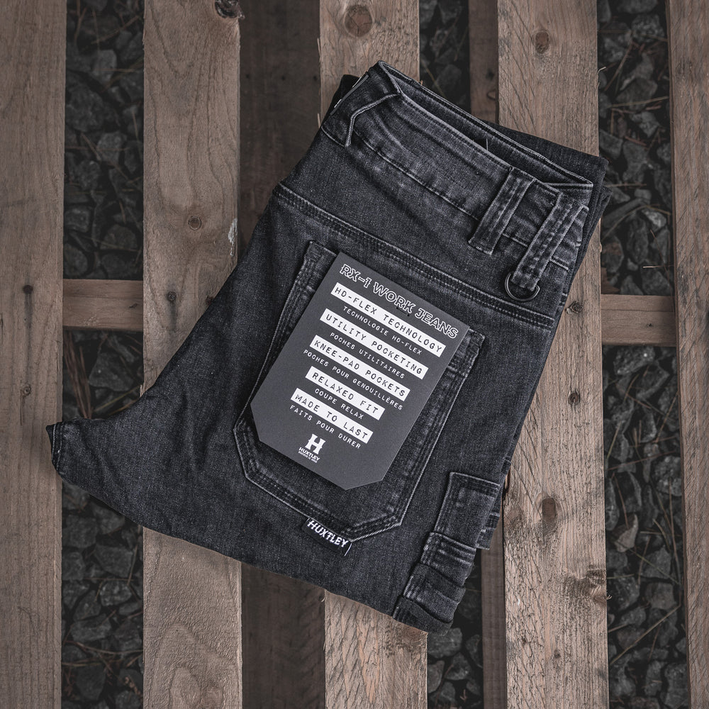 
                  
                    Folded RX-1 Relaxed Work Jeans in Stonewashed Black displayed on a wood pallet. Features utility pocketing, reinforced knees, and Huxtley’s HD-FLEX tech for next-level mobility and durability.
                  
                