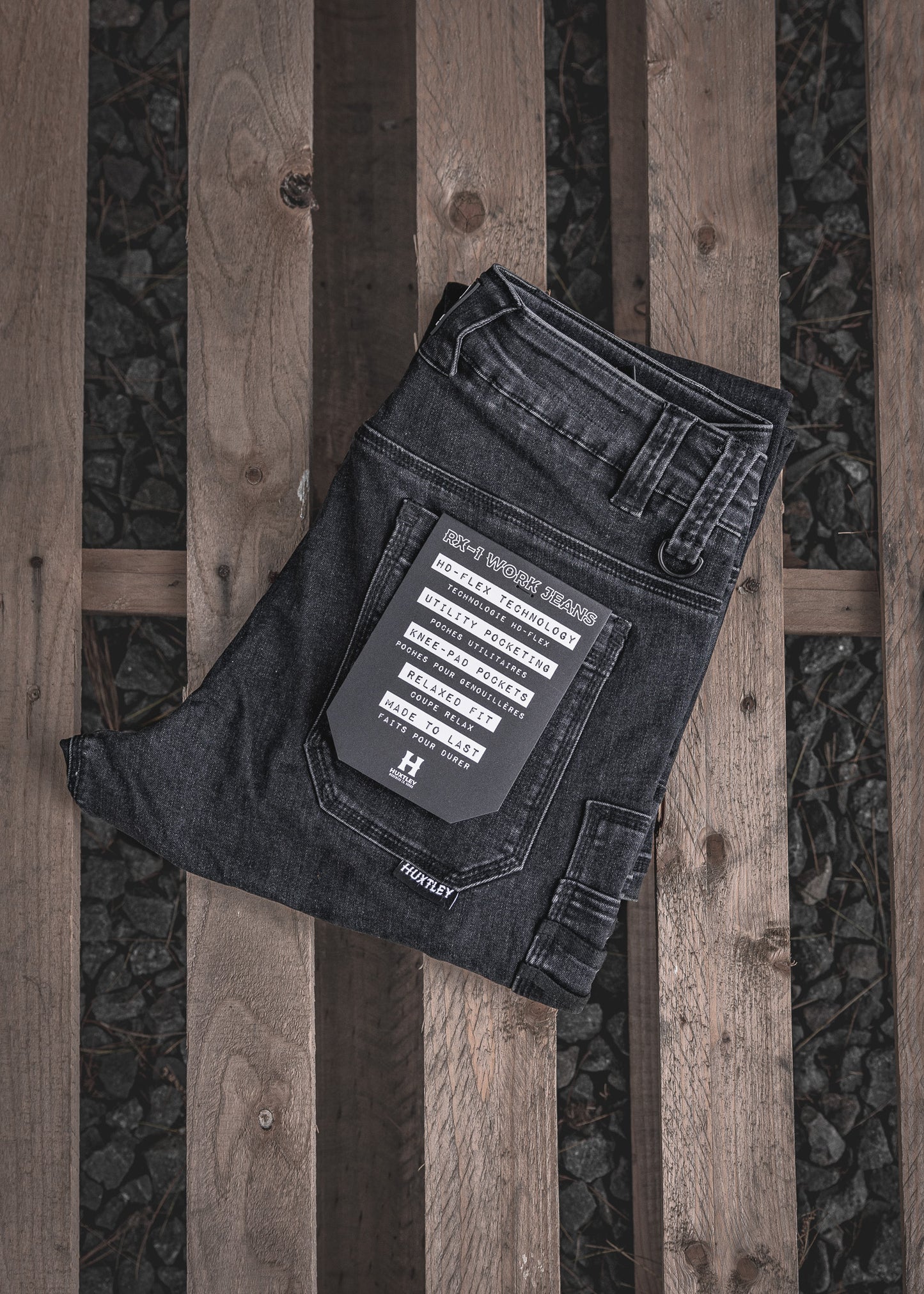 
                  
                    Folded RX-1 Relaxed Work Jeans in Stonewashed Black displayed on a wood pallet. Features utility pocketing, reinforced knees, and Huxtley’s HD-FLEX tech for next-level mobility and durability.
                  
                